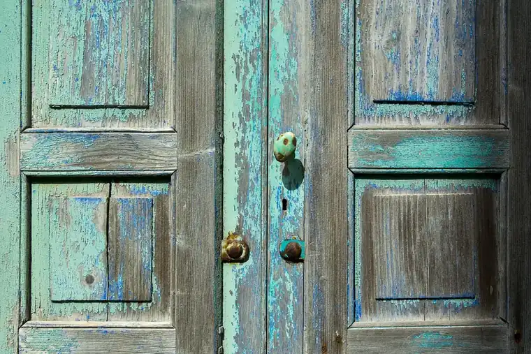 Close-up of weathered panelled timber doors with flaking blue and green paint and an old round knob