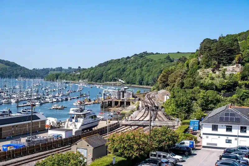 Marina full of moored boats beside a riverside railway line, with wooded hills rising on the far bank