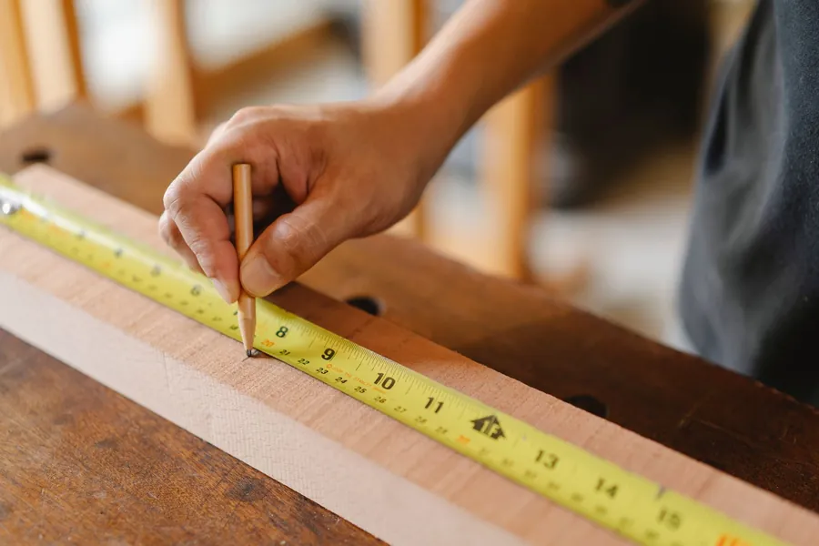 Joiner's hand marking a length of timber with a pencil against a yellow tape measure