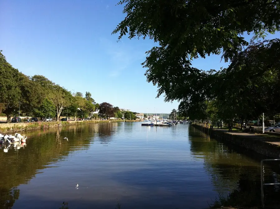 Tree-lined tidal creek with a stone quay wall and small boats moored at the far end