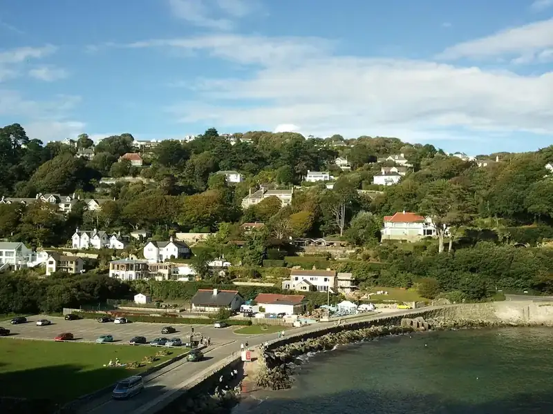 White houses scattered over a wooded hillside above a small sandy cove and a seafront car park