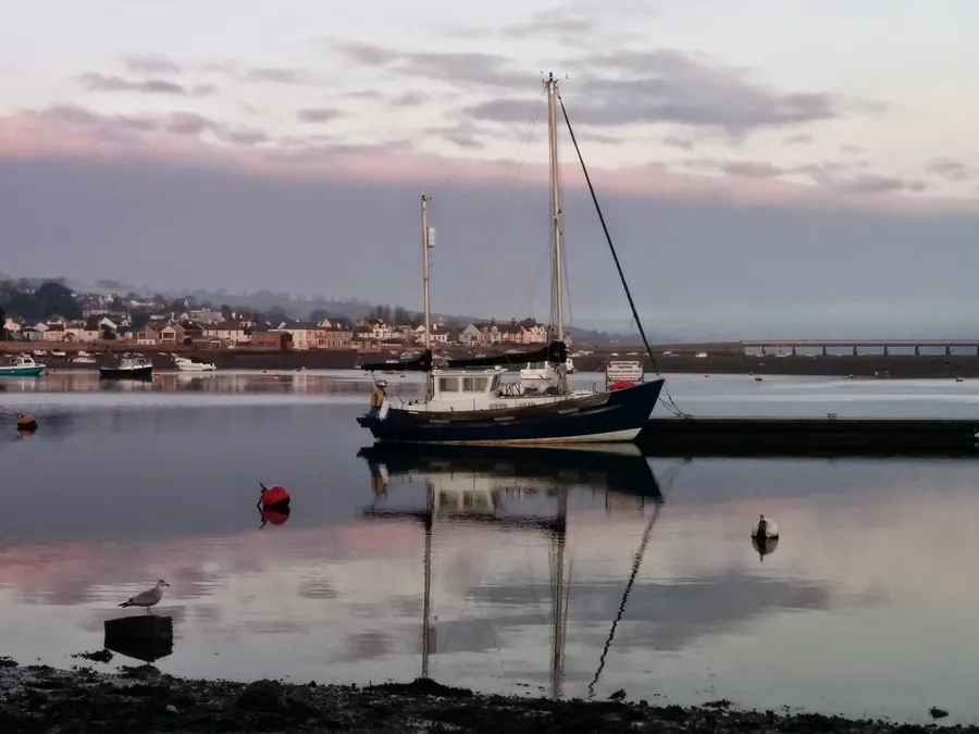 Sailing boat moored on a still estuary at dusk, with houses along the far shore
