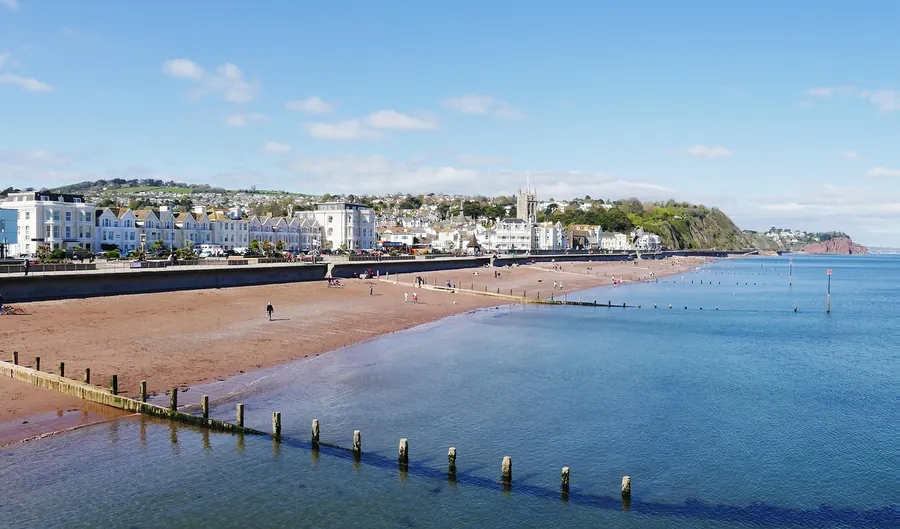 Row of pale painted terraces behind a long red-sand beach and wooden groynes on a sunny day