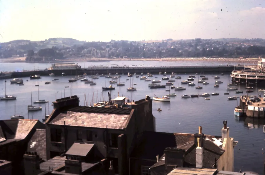 Older photograph looking over Torquay harbour to the wooded hillside beyond, where Victorian villas with sash and bay windows climb above the seafront