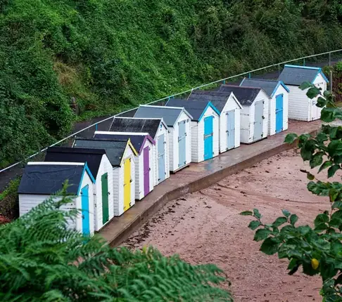 A row of colourful beach huts beneath a green wooded slope
