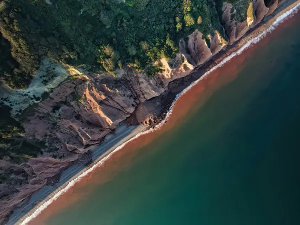 Red earth cliffs and woodland running down to a narrow strip of beach, from the air