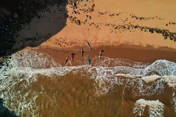 People walking on a red sand beach at the edge of the waves, seen from above