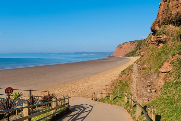 Red cliffs above a long sandy beach, with a path and railings on the hillside