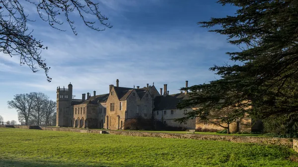 A castellated stone manor house beyond an open field and trees