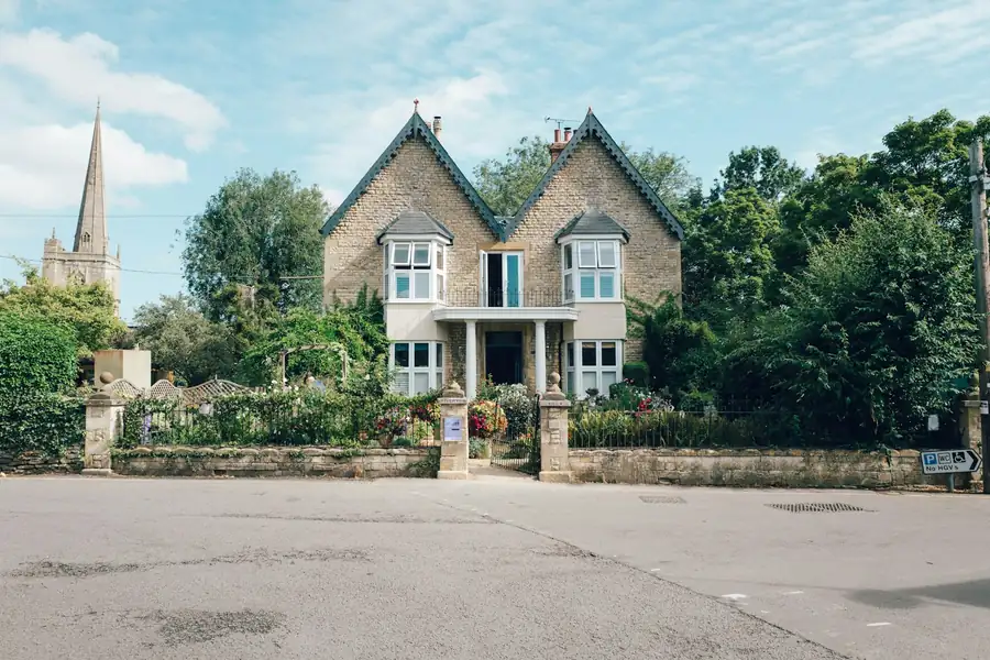 A gabled stone villa behind a garden wall, with a church spire beyond