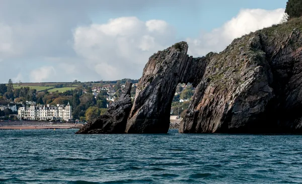 A natural rock arch rising out of the sea below a wooded clifftop