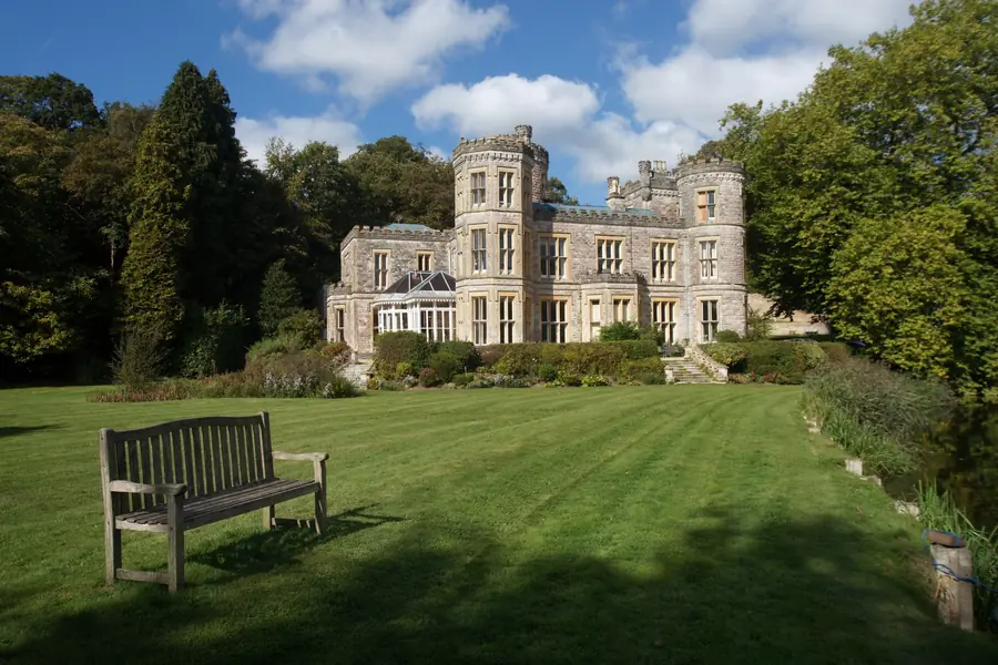 A grey stone villa with tall windows above a sloping lawn and a bench