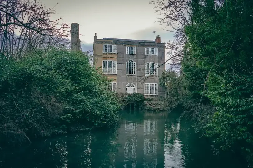 A tall stone house half hidden among dense trees beside water